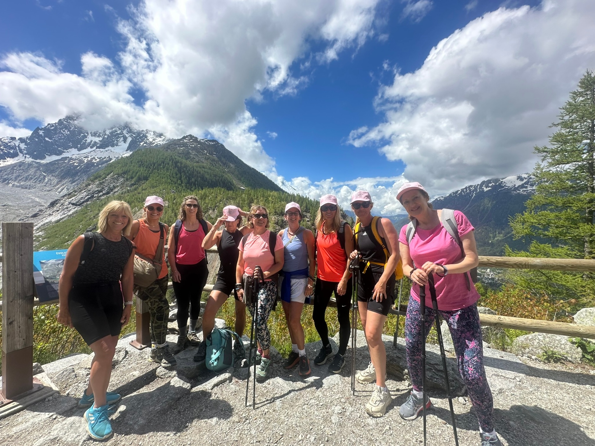 Laura leading a happy hiking group of women in matching pink caps, holding hiking poles on a stunning mountain trail in Chamonix under a blue sky with fluffy white clouds