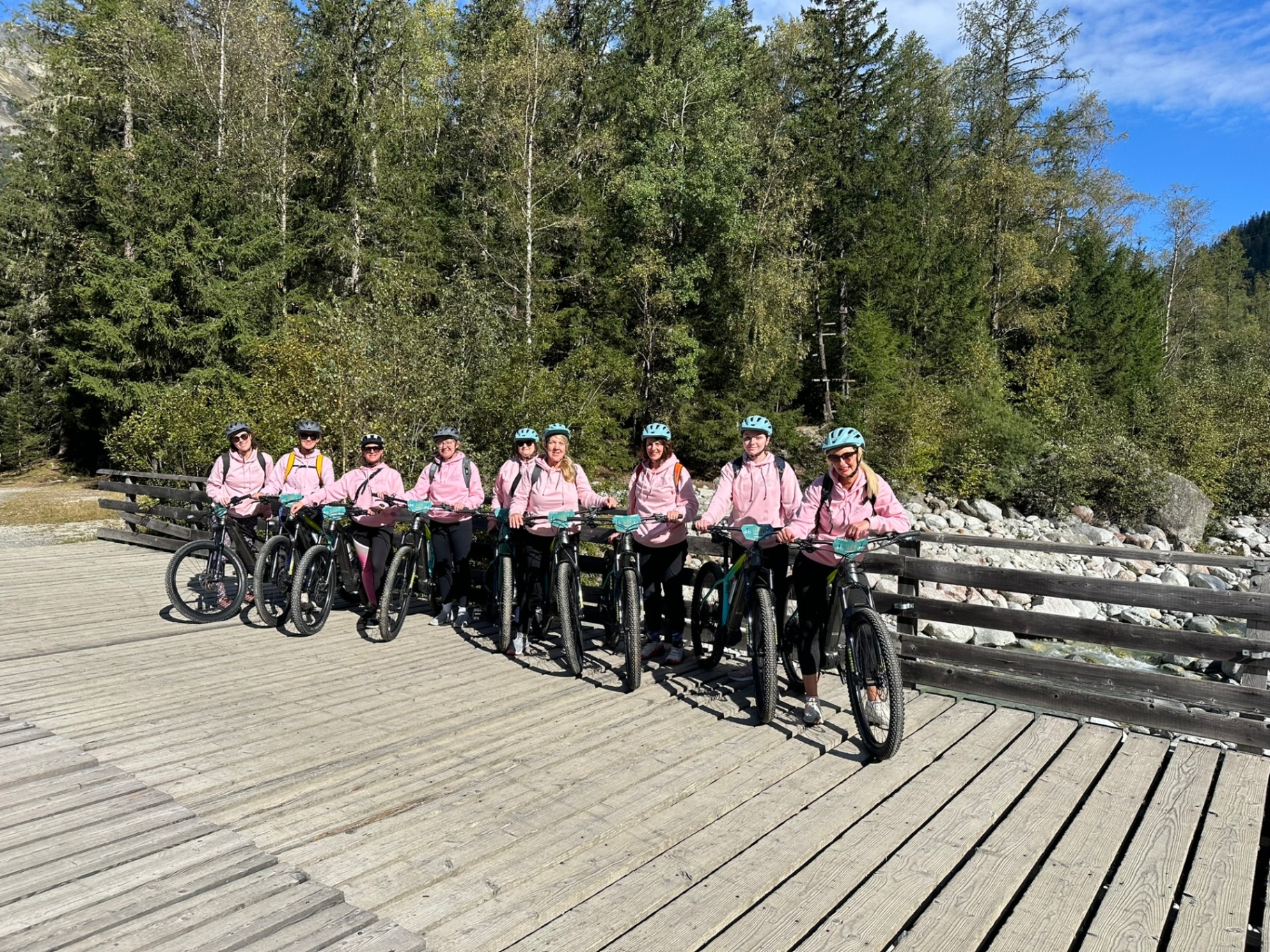 Group of women in matching pink hoodies and turquoise helmets standing with e-bikes on a wooden bridge, enjoying a summer retreat mountain biking adventure in Chamonix